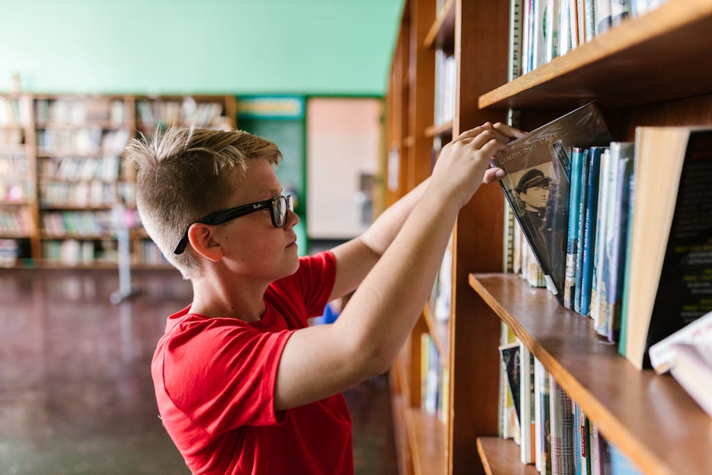 A young boy wearing glasses selects a book from a library bookshelf, emphasizing education and reading.
